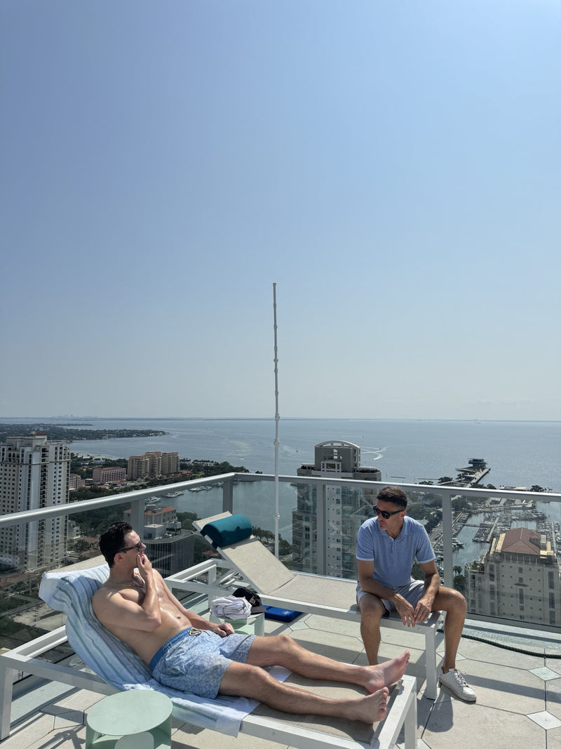 Two men relaxing on a high-rise luxury terrace with a waterfront city view and MR.PATIOMISTER