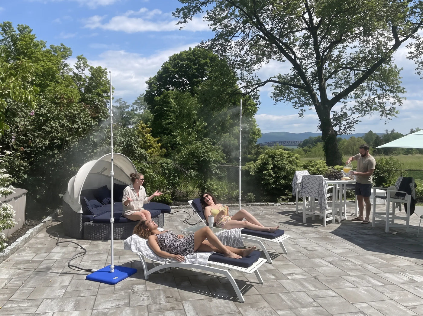 Three friends laughing on a sunny patio while two Mr Patio Mister misting systems run beside them, with a man pouring drinks in the background — portable patio misting system cooling an outdoor patio